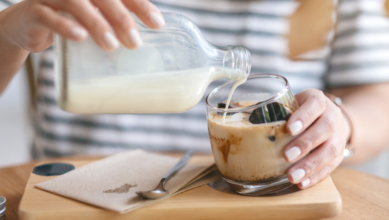 Person gießt Milch aus einer Glasflasche in ein Glas mit Eiskaffee auf einem Holztablett.