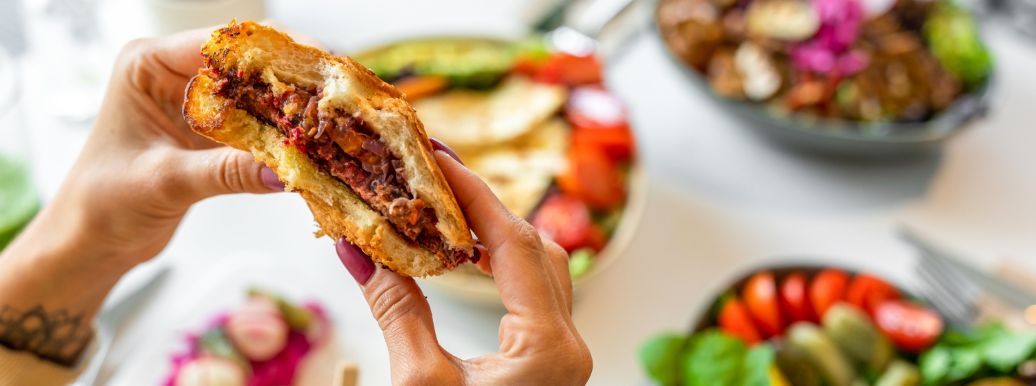 Nahaufnahme von Händen, die einen herzhaften Burger halten, umgeben von bunten Beilagen.