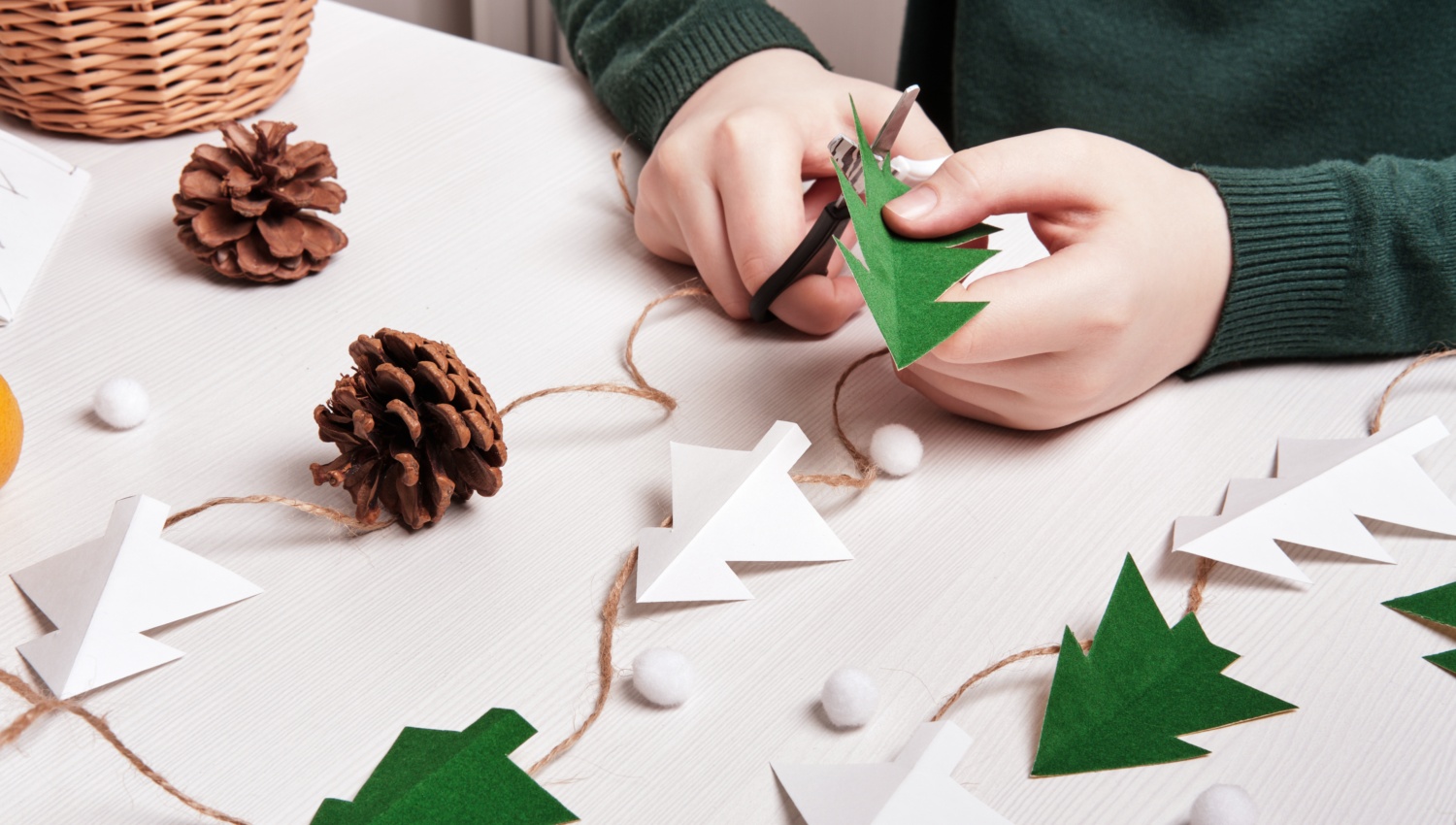 Hände basteln eine Weihnachtsgirlande mit grünen und weißen Papierbäumen, Tannenzapfen und Filzkugeln.
