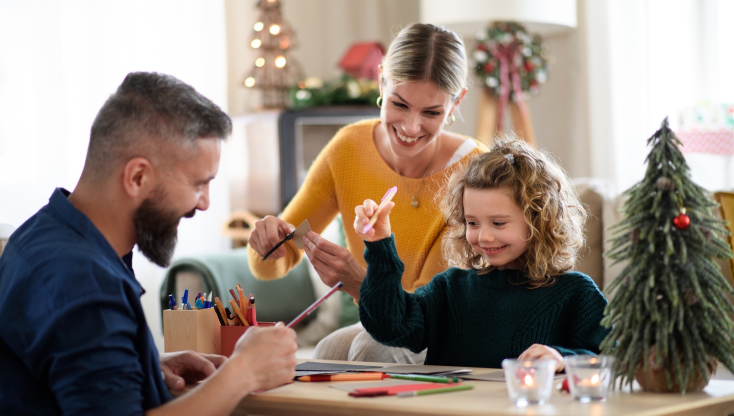 Familie bastelt fröhlich Weihnachtskarten am Tisch mit Bastelmaterialien und Weihnachtsbaum im Hintergrund.