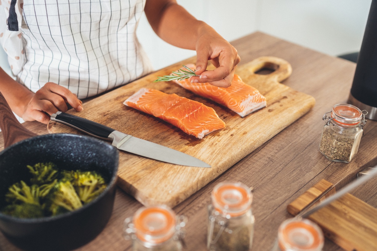 Eine Person bereitet zwei Lachsfilets auf einem Küchenbrett zu. Daneben frische Gewürze und ein Kochtopf.
