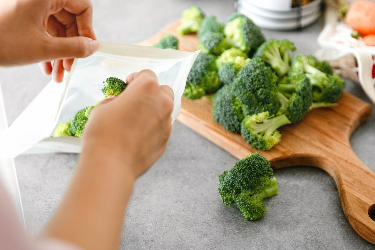 Person legt Brokkoliröschen, die auf einem Holbrett liegen in einen Gefrierbeutel.
