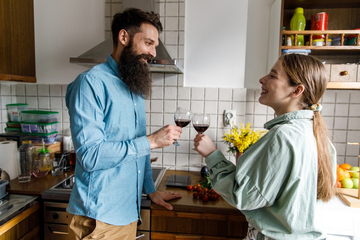 Mann und Frau in einer Küche schauen sich lächelnd in die Augen und stoßen mit Rotweingläsern an.