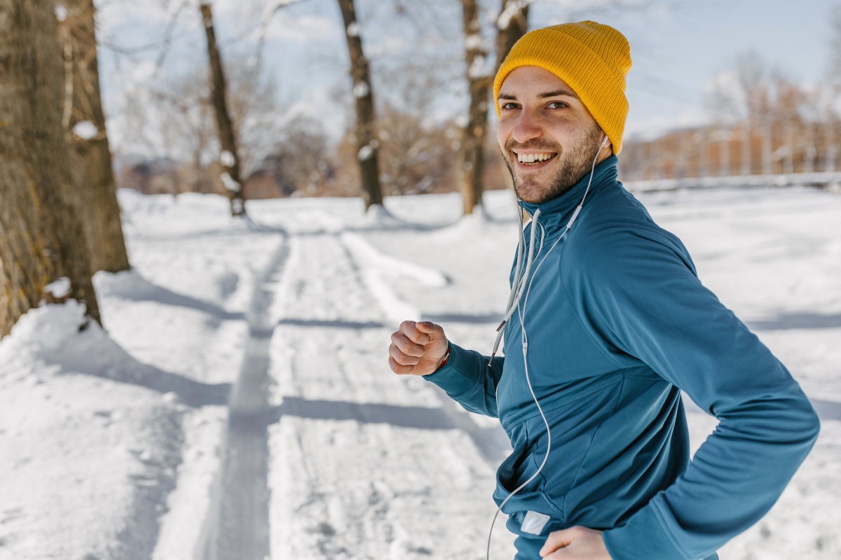 Ein Mann mit Sportkleidung und Mütze joggt lächelnd auf schneebedeckter Laufstrecke im Winter.
