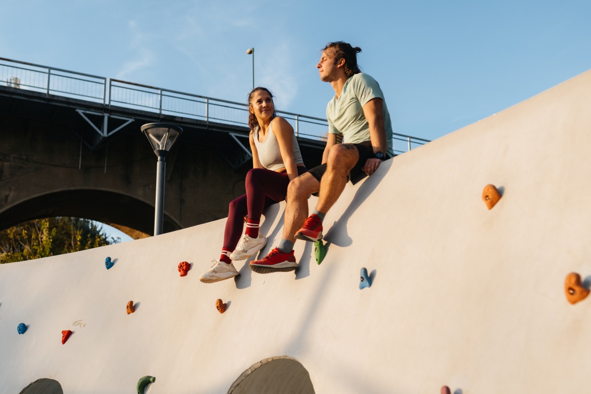 Zwei junge Erwachsene sitzen auf einer Kletterwand im Freien, lächeln und genießen das Sonnenlicht, mit Brücke im Hintergrund.