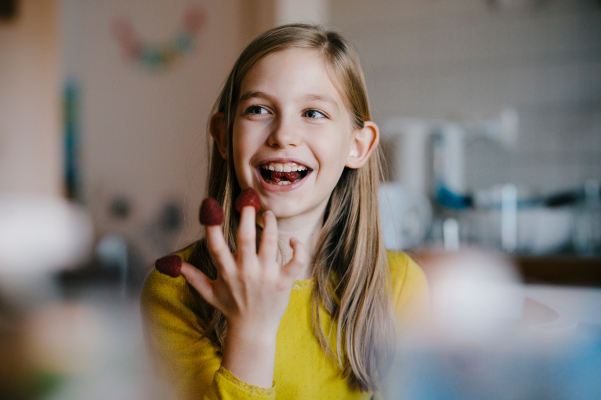 Mädchen isst lachend Himbeeren, die auf ihren Fingern stecken. Freude an gesunden Snacks in der Kinderernährung.