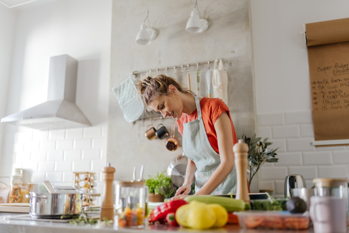 Junge Frau mit Kochschürze  in großer moderner Küche schneidet Gemüse, umgeben von frischen Zutaten.