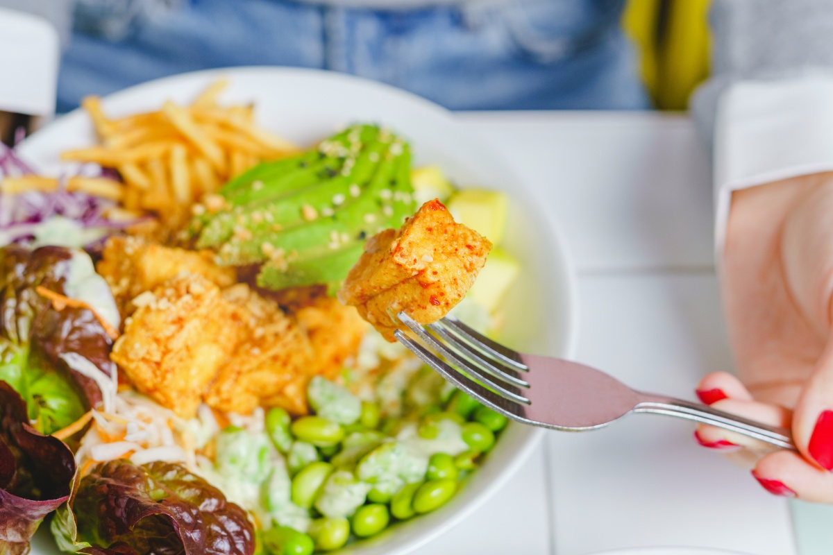 Gabel mit Tofu-Stück über einer farbenfrohen Salatschüssel mit Avocado, Edamame, Pommes und Blattsalat.