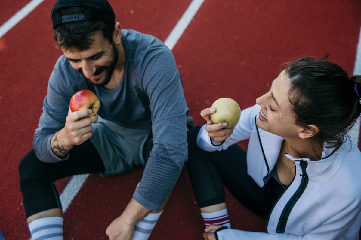 Zwei Personen in Sportkleidung sitzen auf einer Laufbahn und essen lächelnd Äpfel nach einem Training.