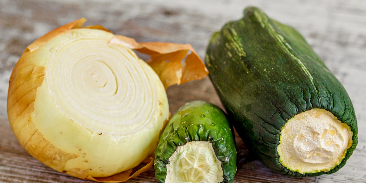 Eine ältere, angeschnittene Zwiebel, Zucchini und Gurke liegen nebeneinander auf einer Küchenplatte.