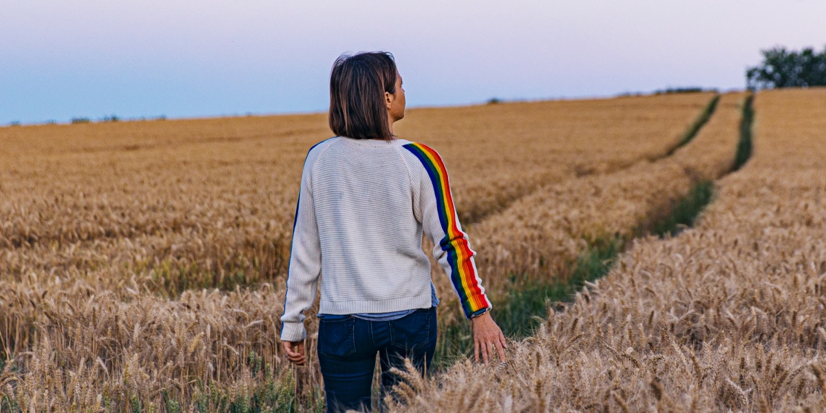 Frau im Pullover mit Regenbogenstreifen steht in einem Getreidefeld bei Sonnenuntergang.