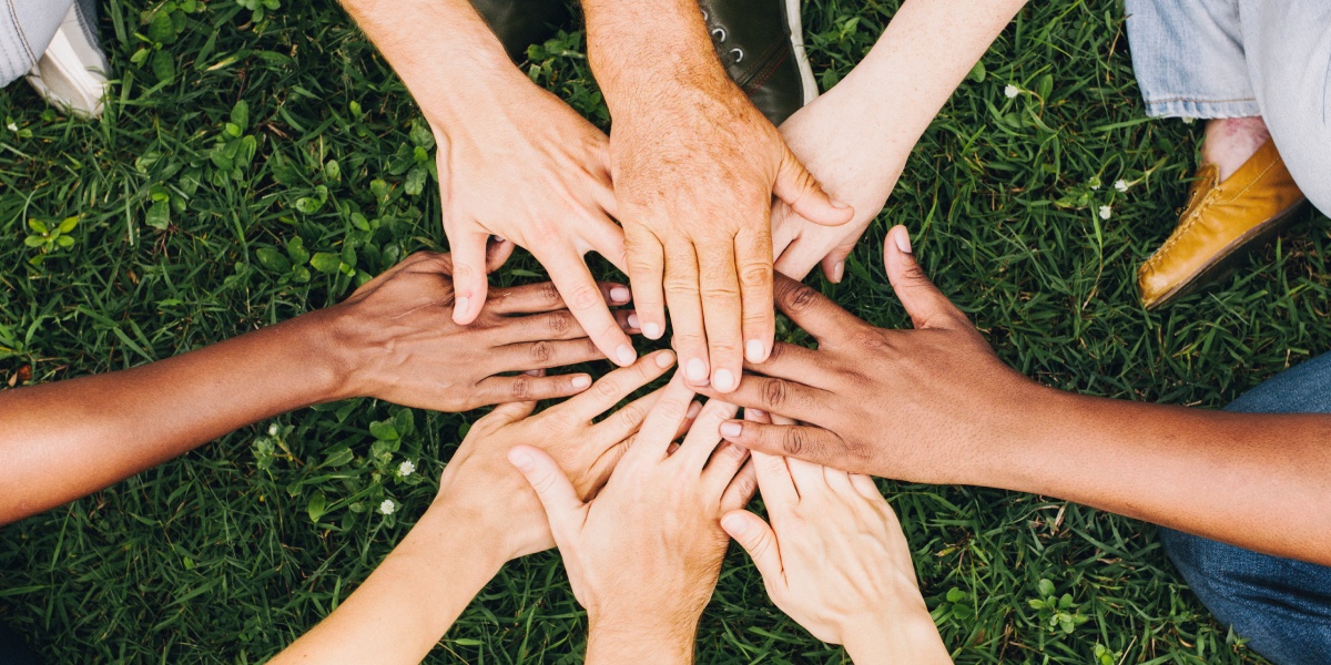 Verschiedene Hände legen sich aufeinander im Kreis auf grünem Gras.