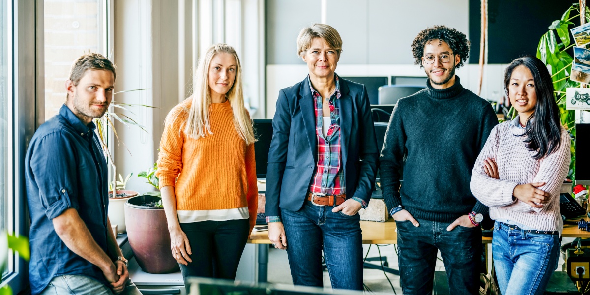 Fünf Teammitglieder in lässiger Kleidung stehen in einem modernen Büro und lächeln in die Kamera.