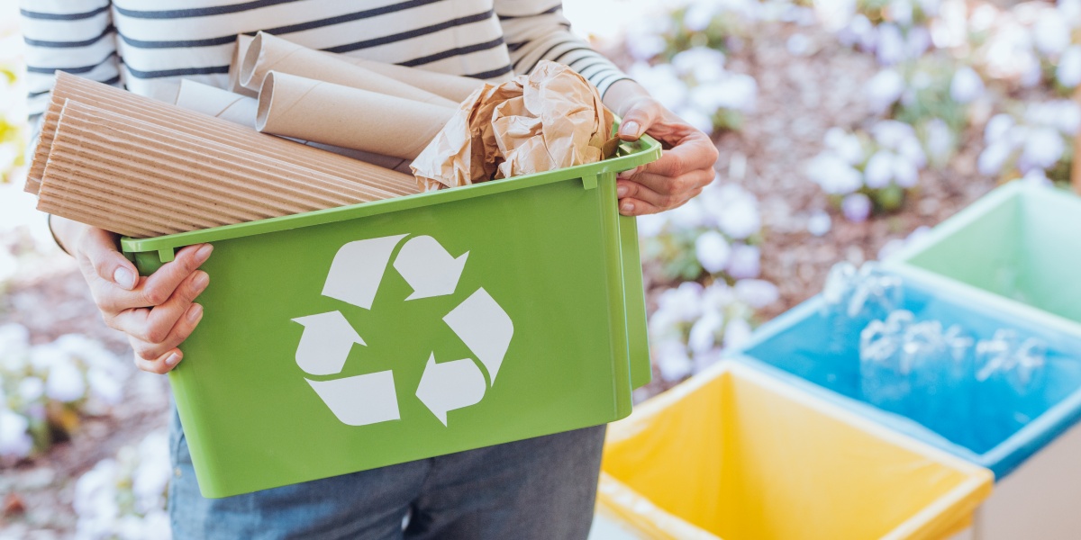 Person trägt Mülleimer mit Papier-Verpackungen, im Hintergrund mehr Behälter zum Mülltrennen für die Kreislaufwirtschaft.