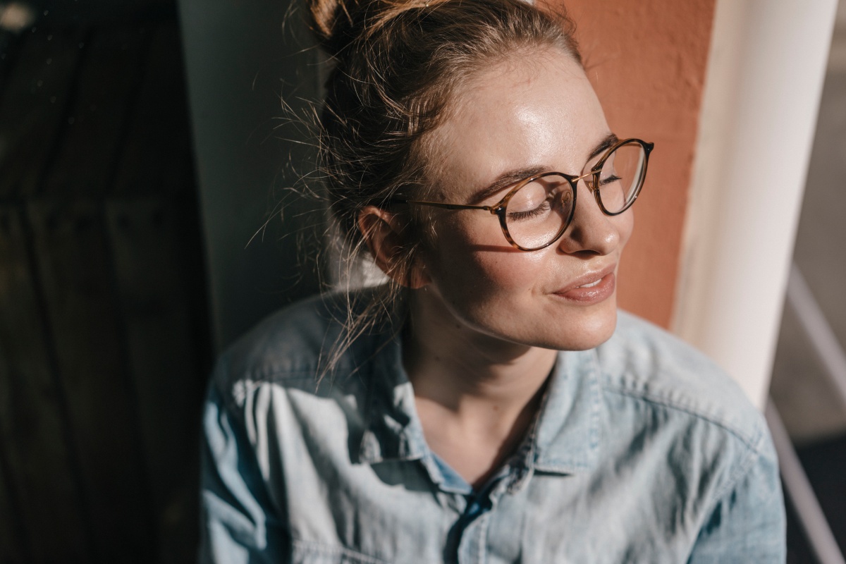 Junge Frau mit Brille schaut mit geschlossenen Augen aus dem Fenster, während die Sonne auf ihr Gesicht scheint.