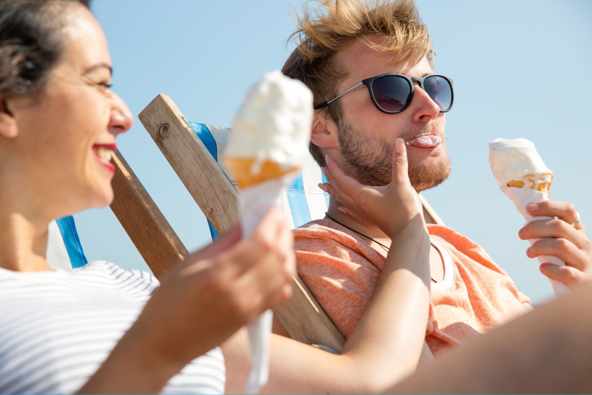Mann und Frau essen Eis am Strand, lachend und entspannt in Liegestühlen unter blauem Himmel.