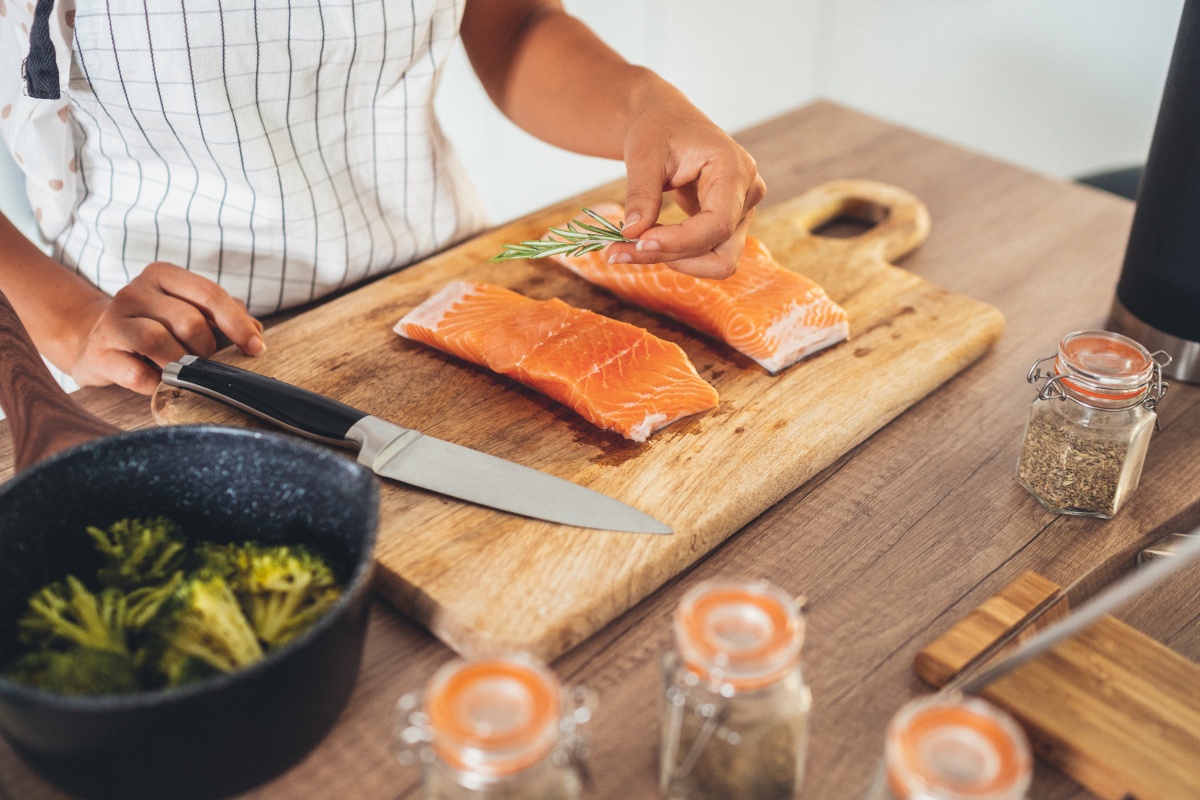 Eine Person bereitet zwei Lachsfilets auf einem Küchenbrett zu. Daneben frische Gewürze und ein Kochtopf.