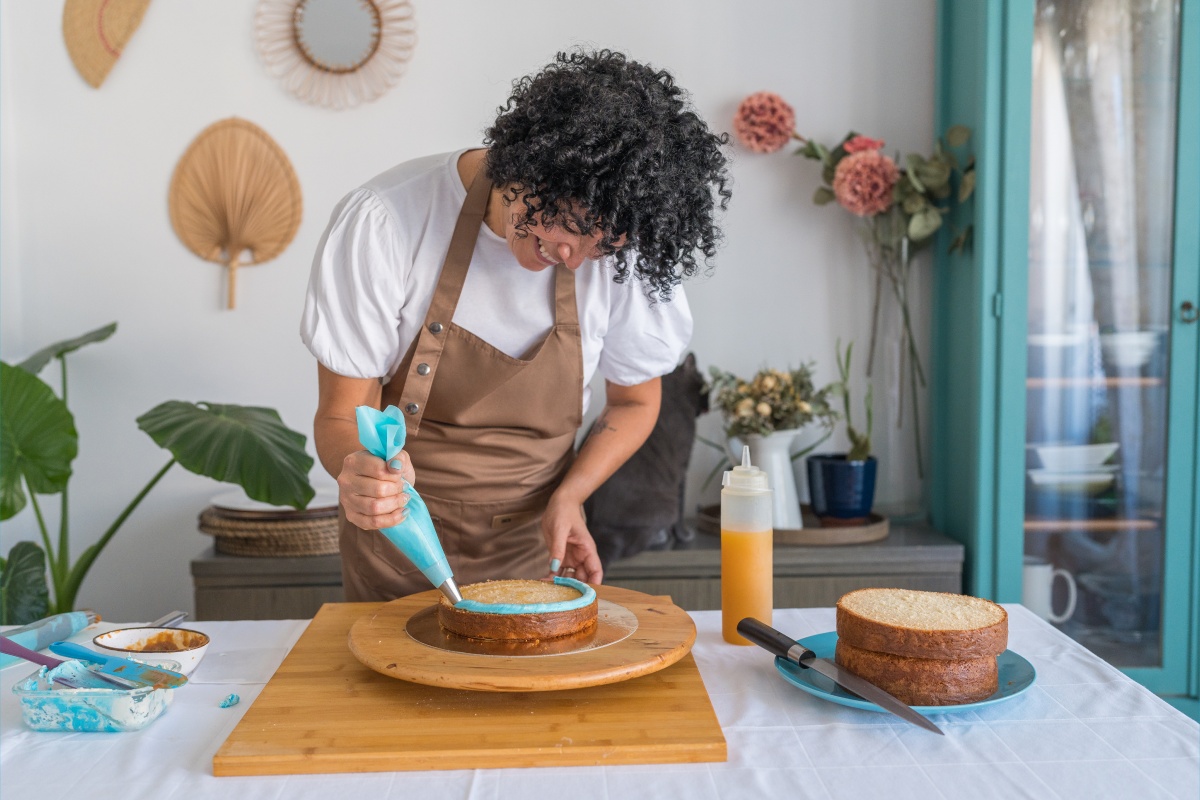 Person in Schürze trägt blaue Glasur auf Kuchen auf, mit Utensilien und zweitem Kuchen auf dem Tisch.