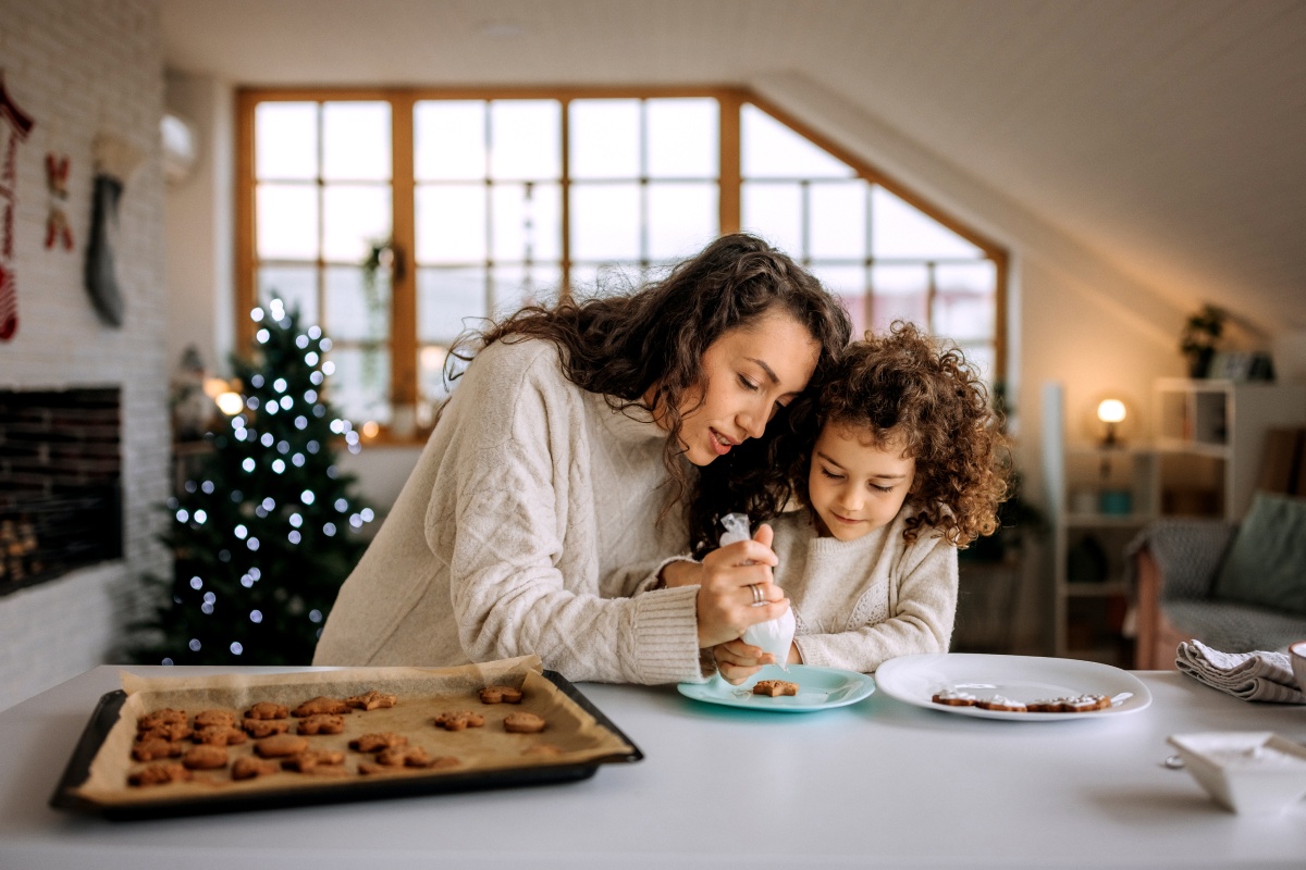 Mutter und Tochter verzieren gemeinsam Kekse in einer weihnachtlich dekorierten Küche, mit Tablett voller frischer Plätzchen.