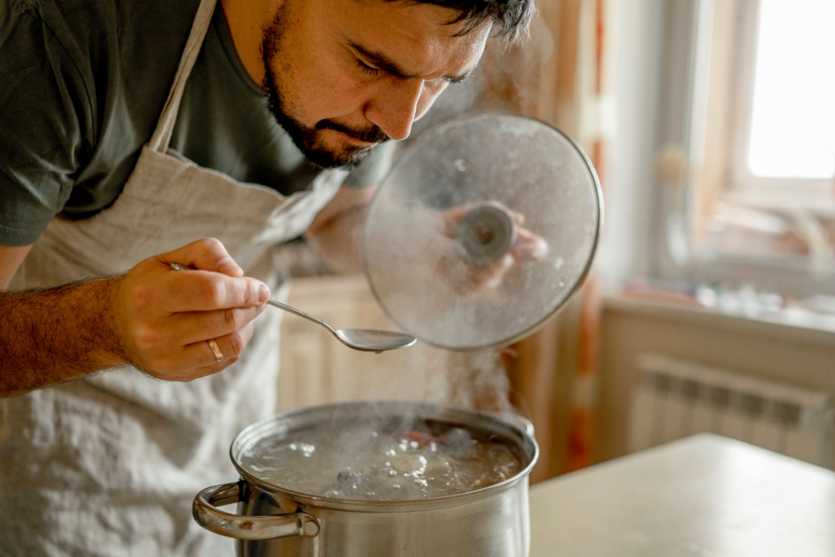 Mann in Schürze probiert Suppe mit einem Löffel aus einem dampfenden Kochtopf.