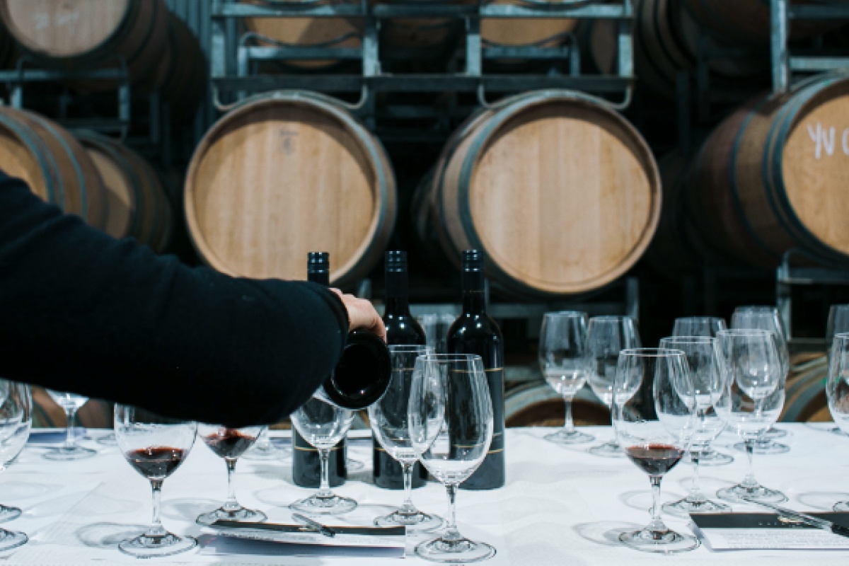 Eine Person schenkt Rotwein in Gläser ein, auf einem Tisch vor gestapelten Holzfässern in einer Weinkellerei.