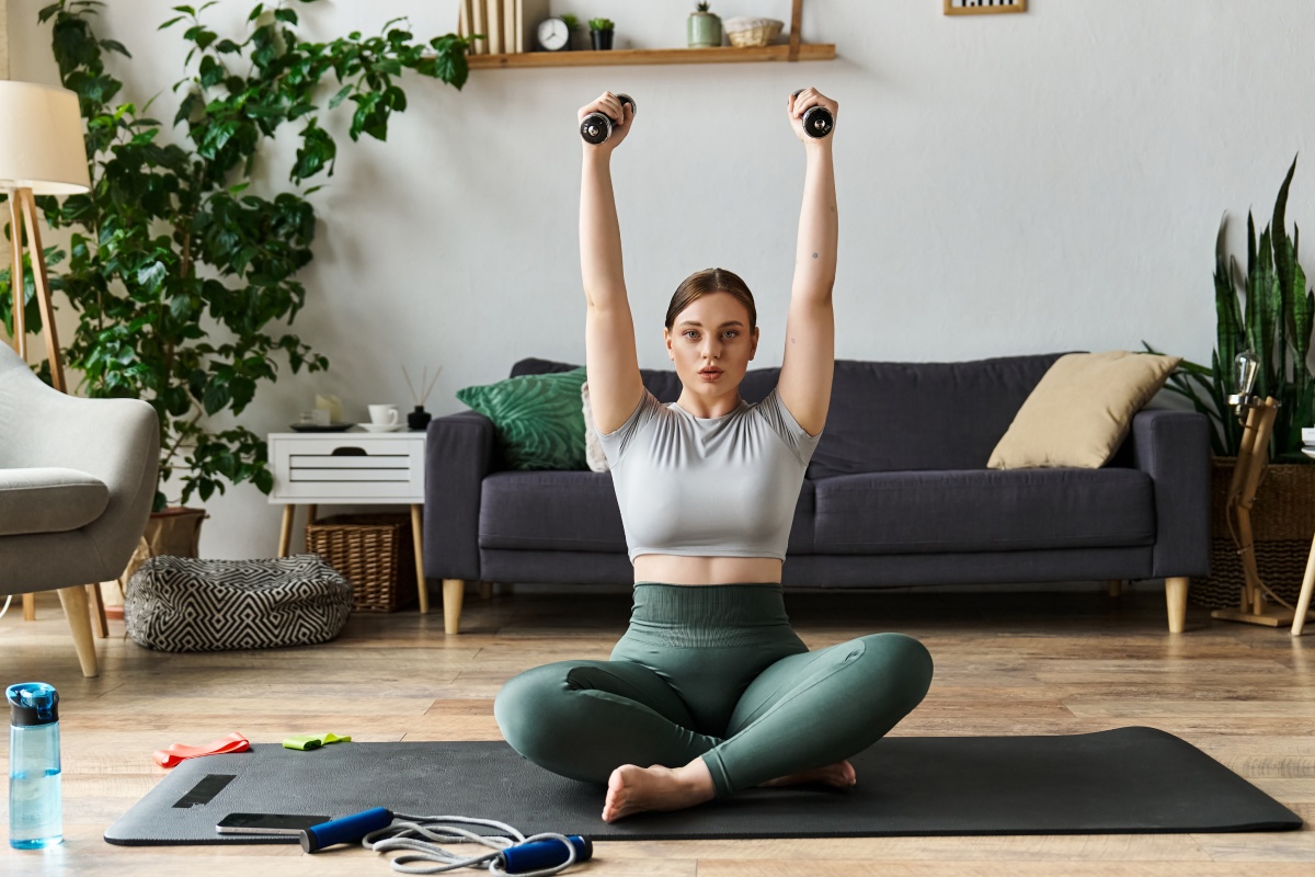 Frau in Sportkleidung hebt Hanteln über den Kopf, sitztend auf einer Yogamatte in einem gemütlichen Wohnzimmer.