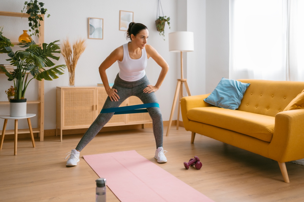 Frau trainiert mit Fitnessband in modernem Wohnzimmer, gelbes Sofa, Pflanzen und rosa Yogamatte im Hintergrund.