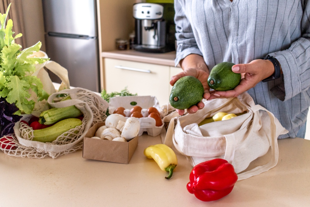 Person hält zwei Avocados über einem Tisch mit frischem Gemüse und Eiern in einer Küche.