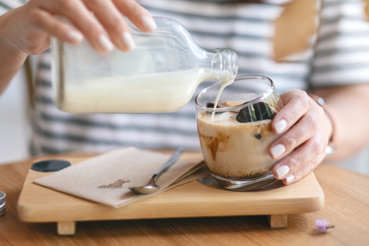Person gießt Milch aus einer Glasflasche in ein Glas mit Eiskaffee auf einem Holztablett.