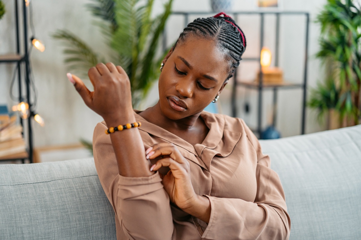 Frau in beiger Bluse sitzt auf dem Sofa und betrachtet ihren Mückenstich auf dem Arm