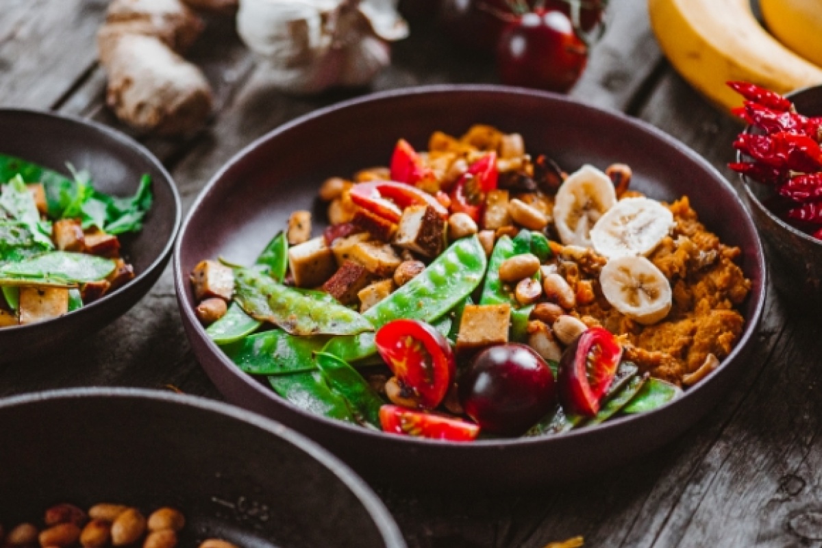 Vegane Buddha-Bowl mit Zuckerschoten, Tomaten, Bananenscheiben, Tofu und Nüssen in einer braunen Schale, rustikal serviert.