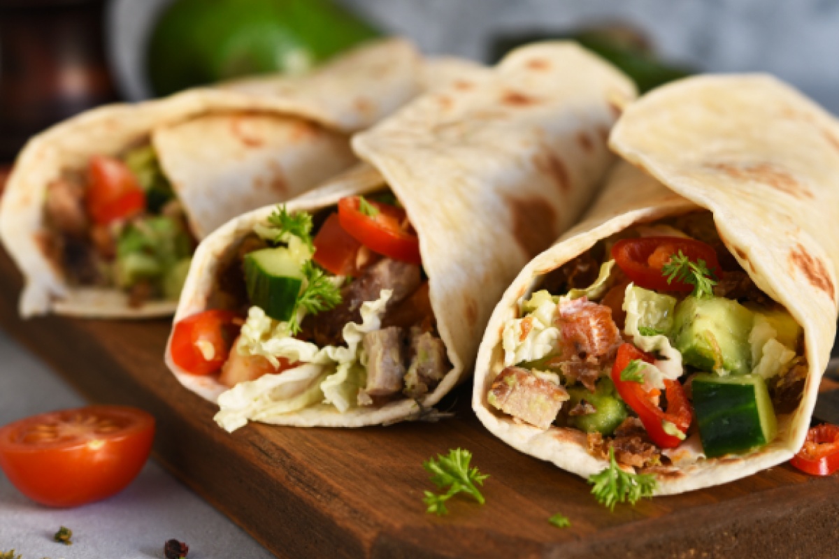 Drei gefüllte Wraps mit frischem Gemüse, Salat, Tomaten und Fleisch auf einem Holzbrett.