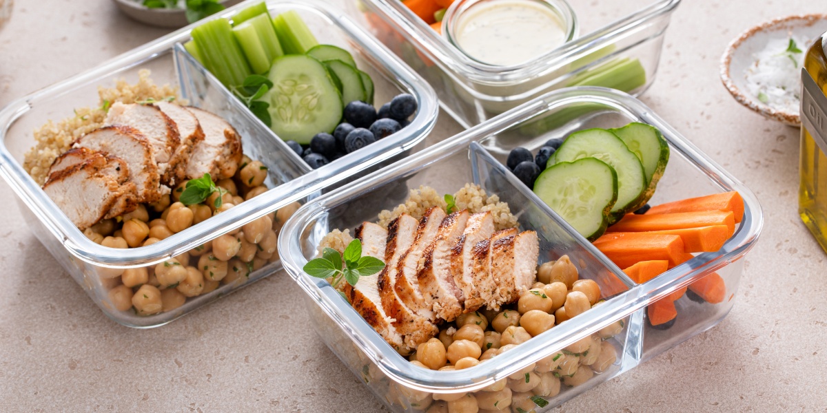 Glasbehälter mit Hähnchenbrust, Kichererbsen, Quinoa, Gurkenscheiben, Blaubeeren, Karotten und Selleriestangen.