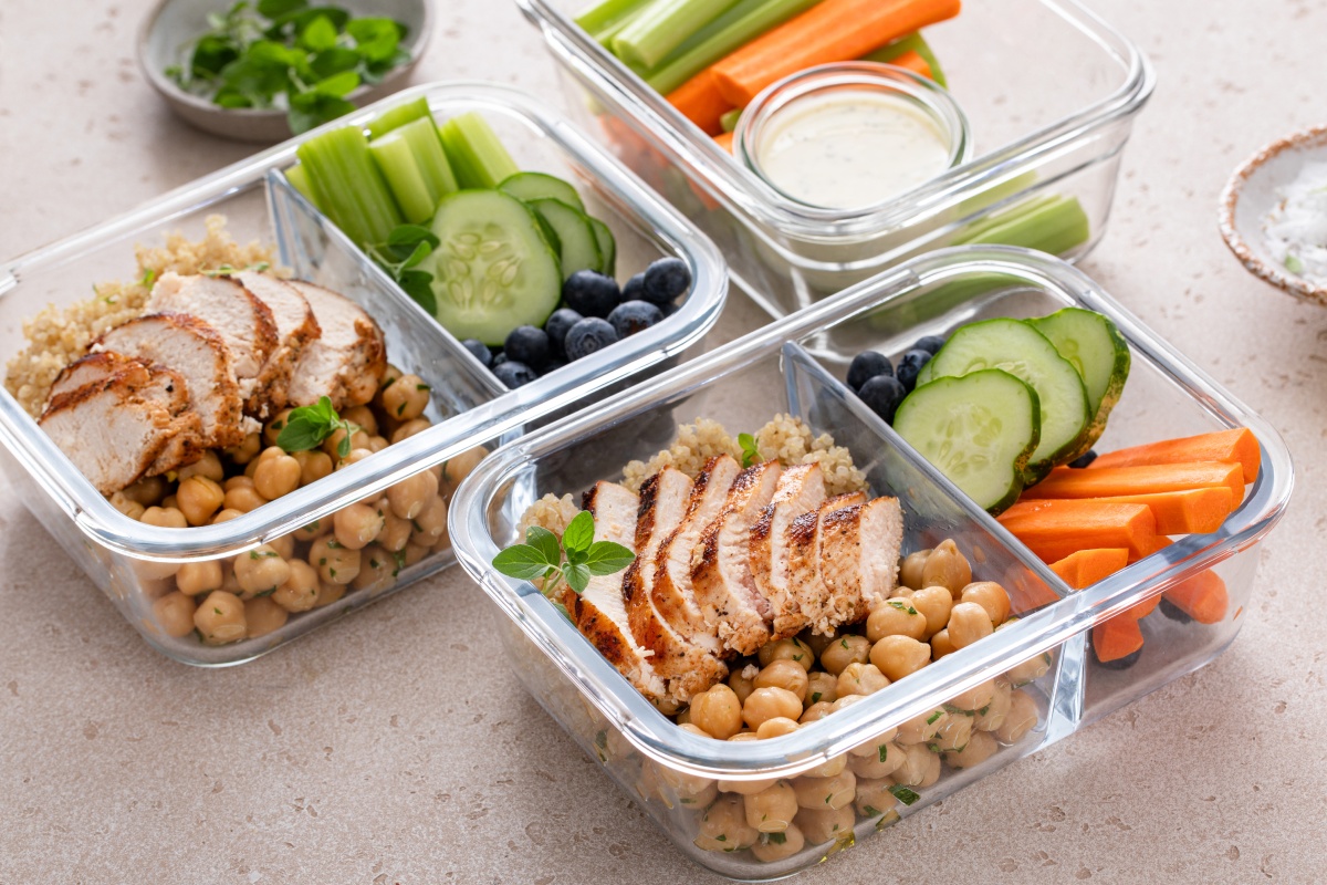 Glasbehälter mit Hähnchenbrust, Kichererbsen, Quinoa, Gurkenscheiben, Blaubeeren, Karotten und Selleriestangen.