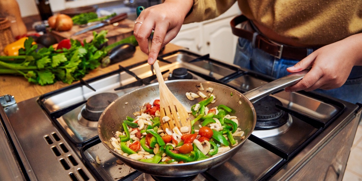 Person brät Gemüse wie Paprika, Tomaten und Champignons in einer Pfanne auf dem Herd.