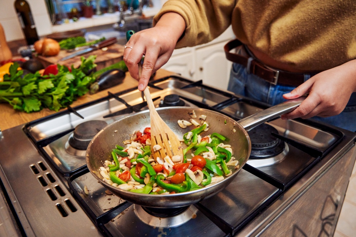 Person brät Gemüse wie Paprika, Tomaten und Champignons in einer Pfanne auf dem Herd.