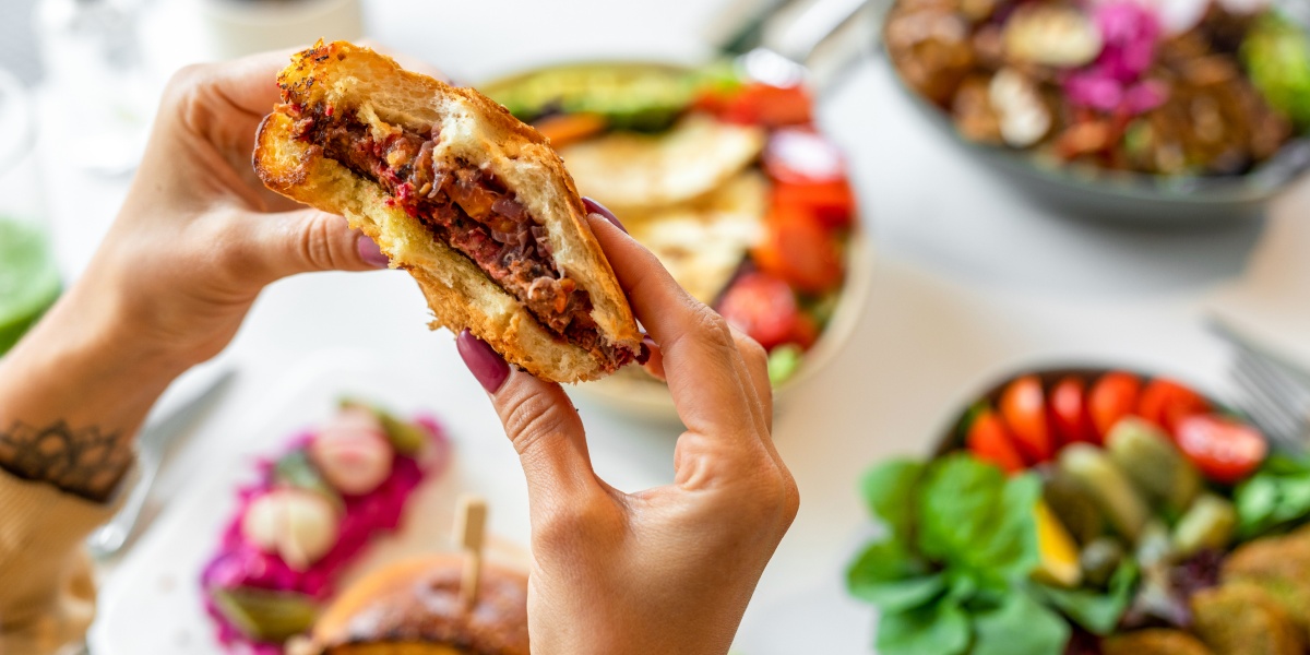 Nahaufnahme von Händen, die einen herzhaften Burger halten, umgeben von frischen Beilagen.