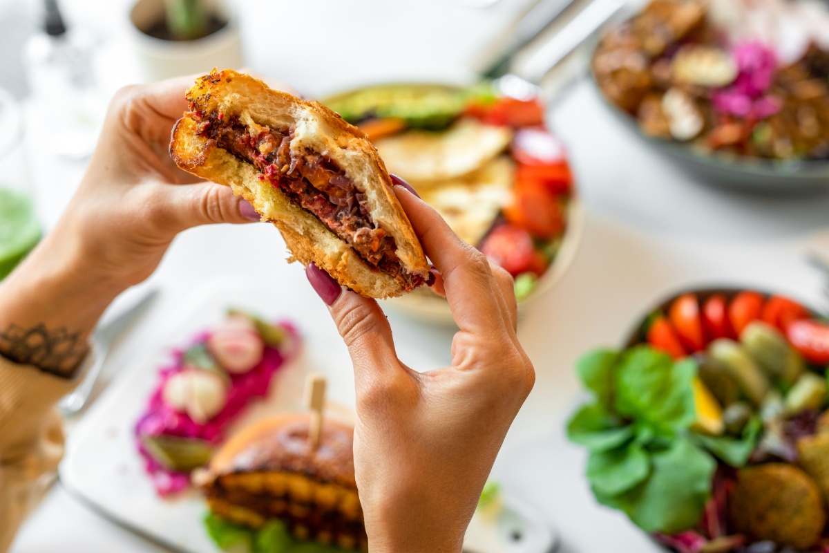 Nahaufnahme von Händen, die einen herzhaften Burger halten, umgeben von frischen Beilagen.