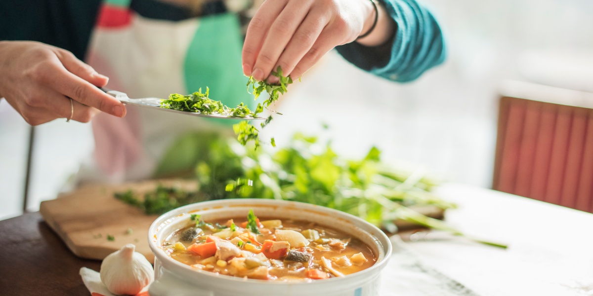Person würzt Gemüsesuppe mit frischen Kräutern, die in den Topf gestreut werden.