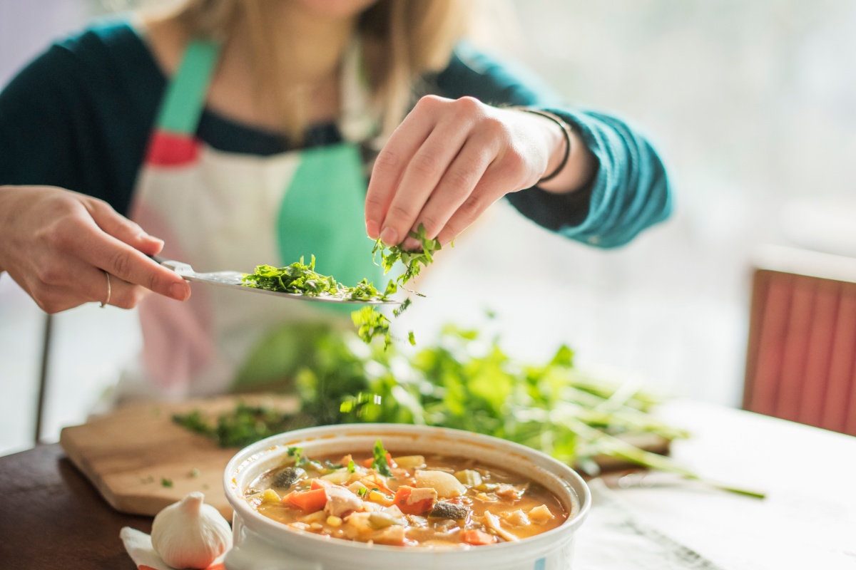 Person würzt Gemüsesuppe mit frischen Kräutern, die in den Topf gestreut werden.
