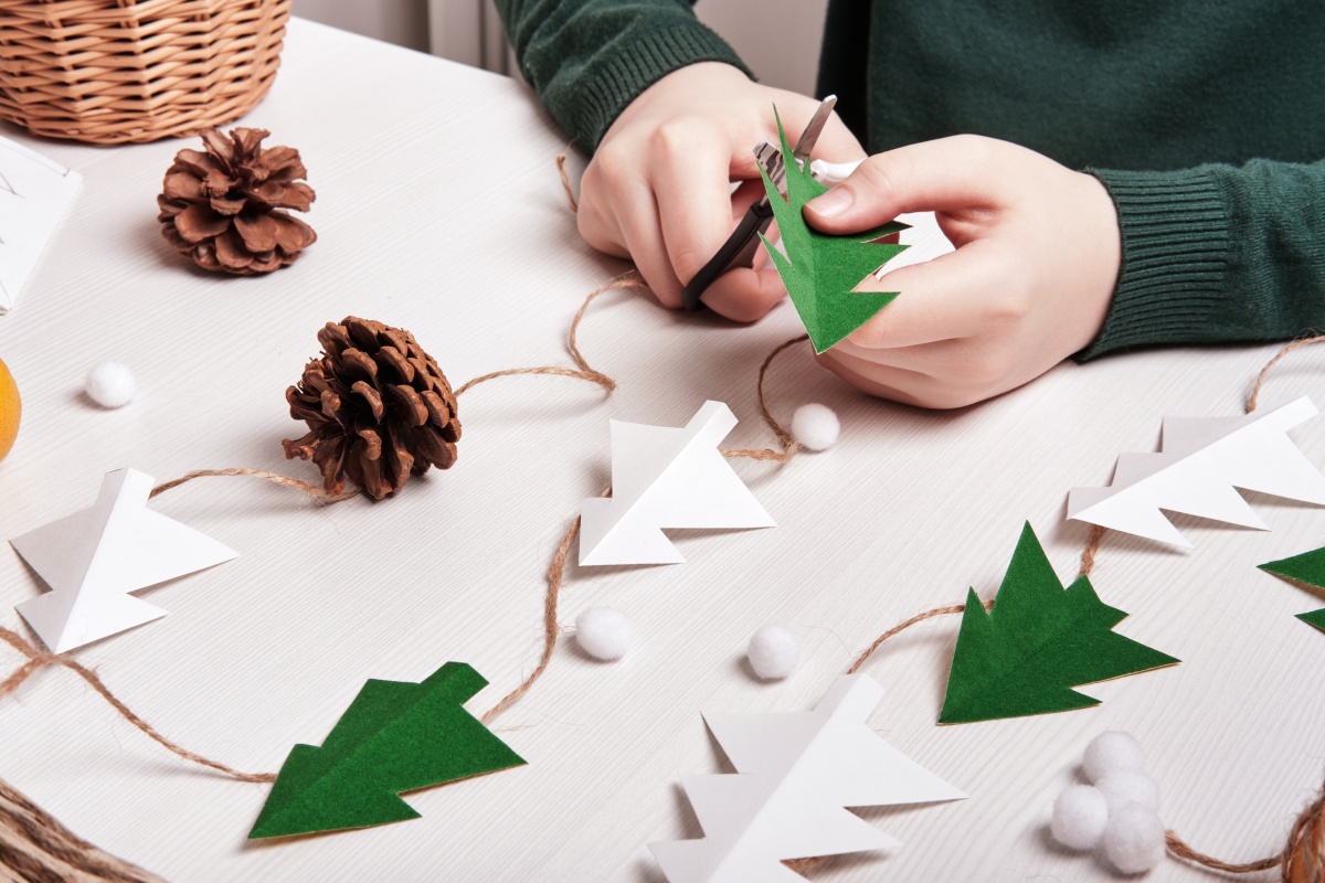 Hände schneiden grüne Papier-Tannenbäume aus, die zusammen mit Zapfen zu Weihnachtsdeko gebastelt werden.