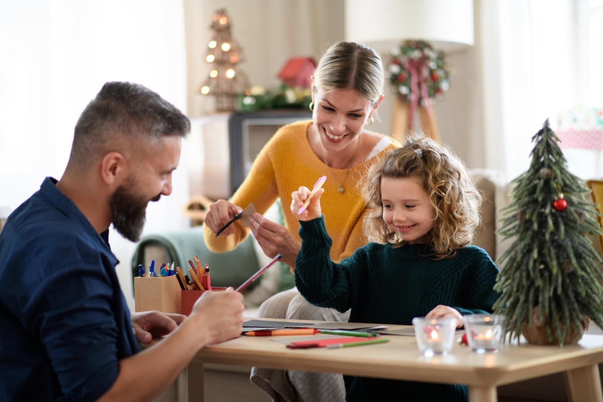 Familie bastelt fröhlich Weihnachtskarten am Tisch mit Bastelmaterialien und Weihnachtsbaum im Hintergrund.