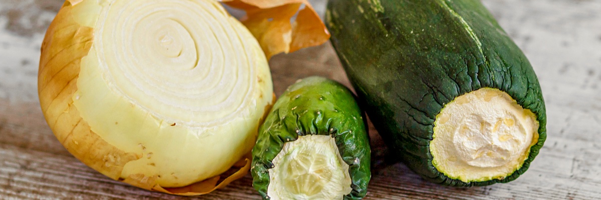 Eine ältere, angeschnittene Zwiebel, Zucchini und Gurke liegen nebeneinander auf einer Küchenplatte.