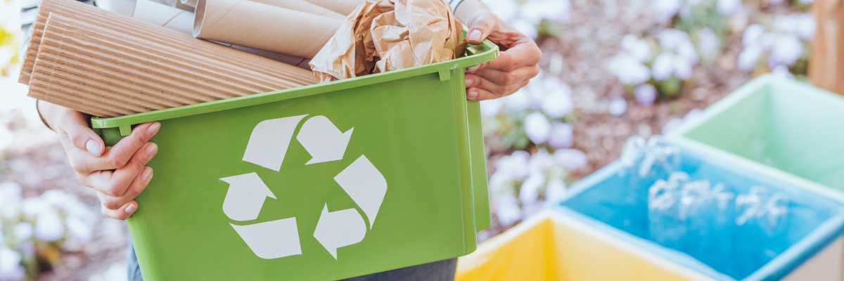 Person trägt Mülleimer mit Papier-Verpackungen, im Hintergrund mehr Behälter zum Mülltrennen für die Kreislaufwirtschaft.