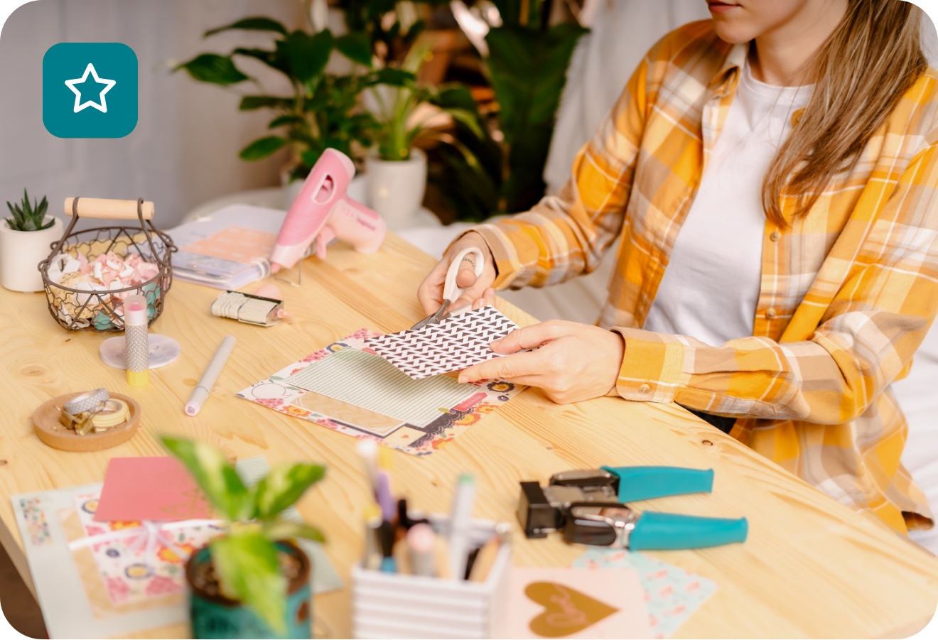 Frau in kariertem Hemd bastelt an einem Schreibtisch mit Papier, Schere und Bastelmaterialien. Kreative DIY-Handarbeitsszene.