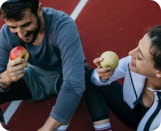 Zwei Personen in Sportkleidung sitzen auf einer Laufbahn und essen lächelnd Äpfel nach einem Training.