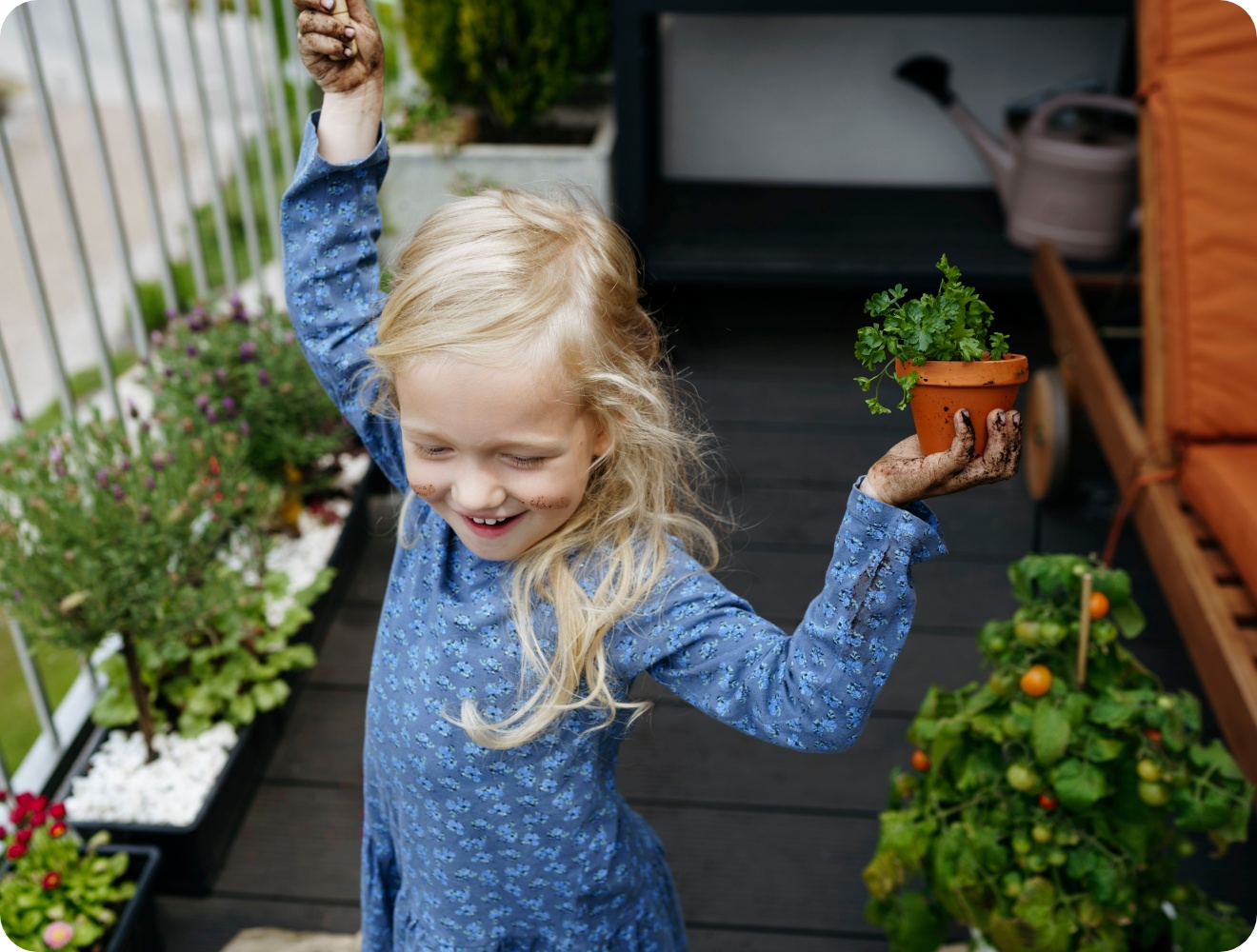 Fröhliches Mädchen mit Erde an den Händen hält Kräutertopf auf Balkon mit Gemüse- und Blumenpflanzen.