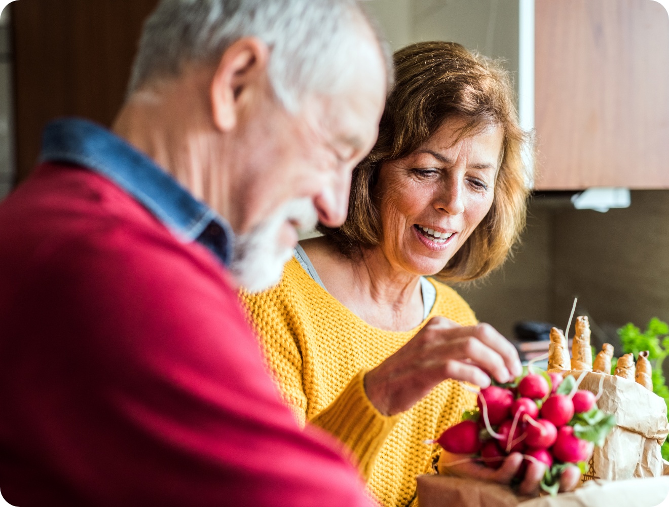 Älteres Paar in der Küche packt frische Radieschen und Karotten aus einer Papiertüte aus.