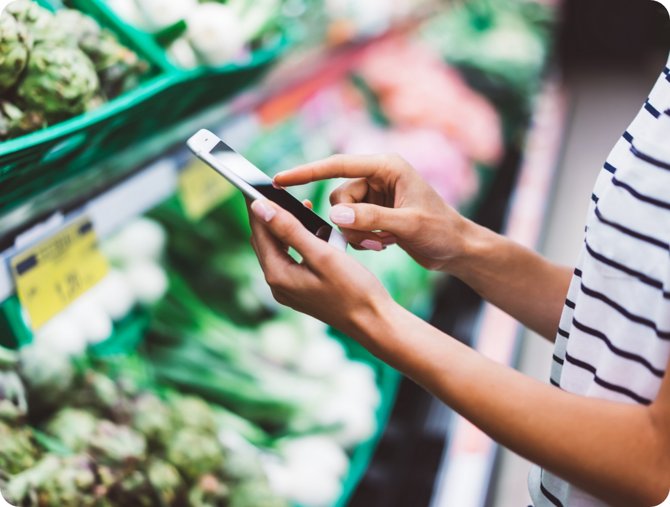Person im Supermarkt tippt auf ein Smartphone, während sie vor einem Regal mit frischem Gemüse steht.