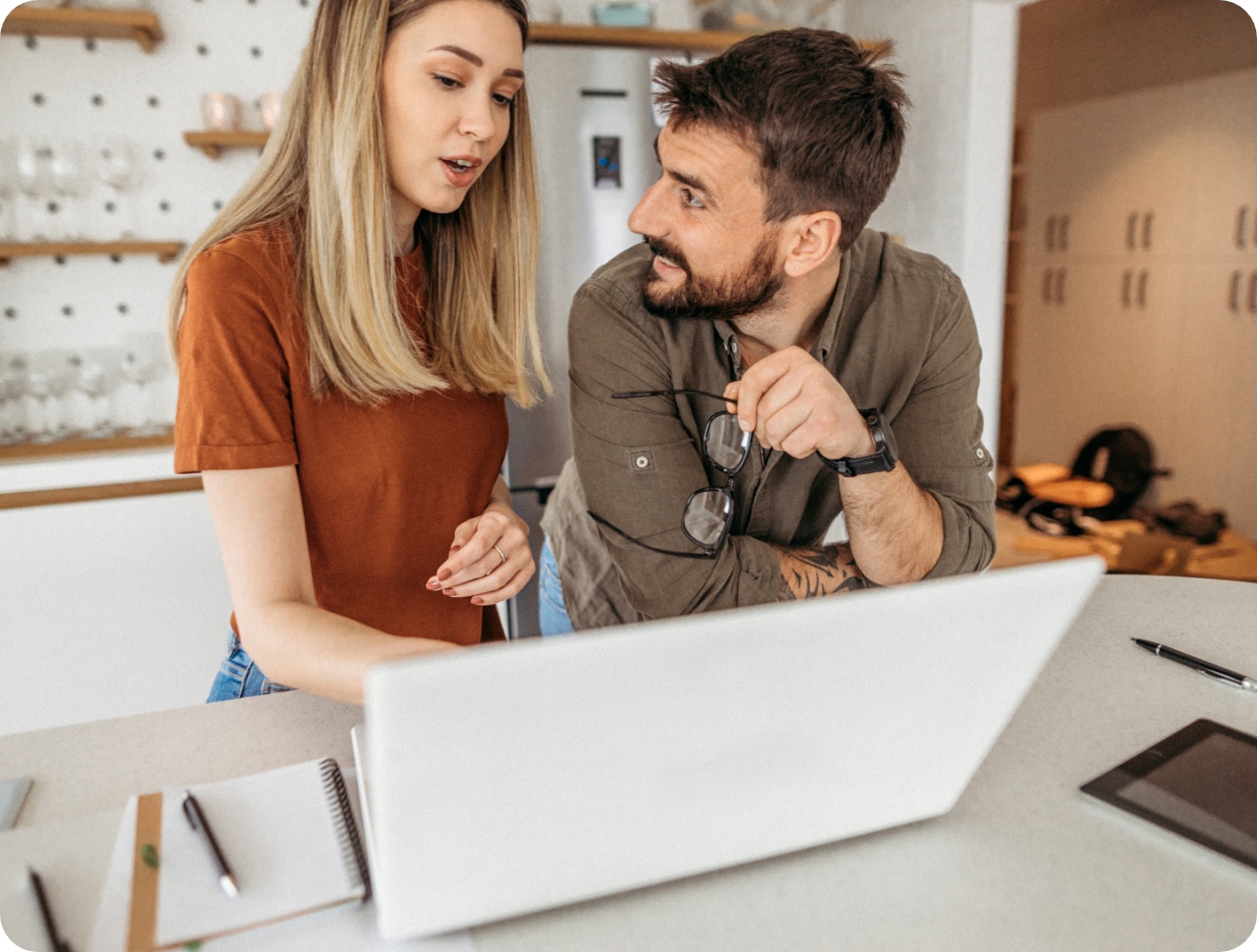 Paar bespricht etwas am Laptop in einer modernen Küche. Notizbuch und Tablet liegen auf dem Tisch.